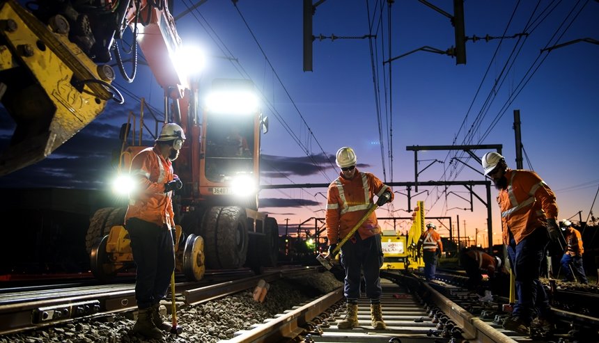 An on the ground view looking at a group of three construction workers laying tracks down on the track line between Sydney Metro's Sydenham and Bankstown Stations  at sundown with the crane digger lights shining bright in the background.