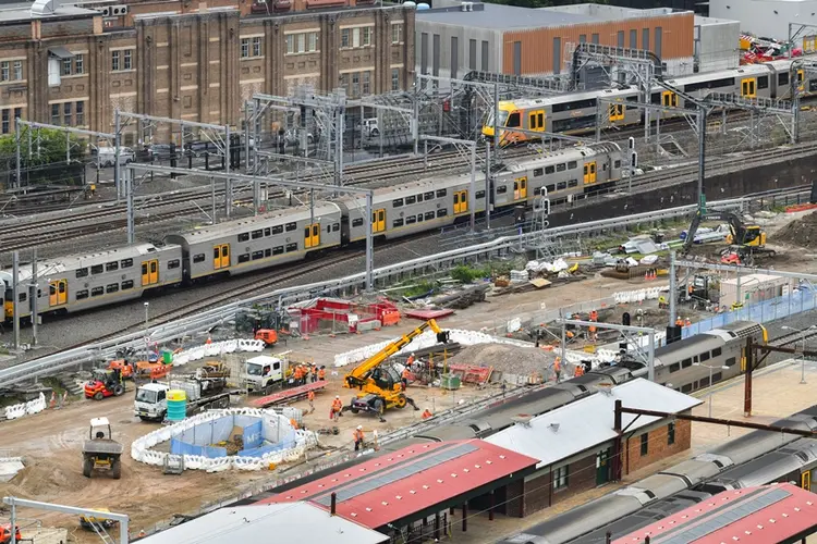 A bird's eye view looking across at the construction site of the Sydney Metro station box at Central Station with many heavy machinery on the ground between the platforms as two trains are leaving the platform in the background.