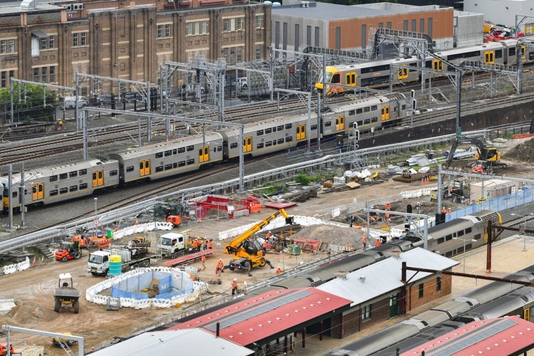 A bird's eye view looking across at the construction site of the Sydney Metro station box at Central Station with many heavy machinery on the ground between the platforms as two trains are leaving the platform in the background.  