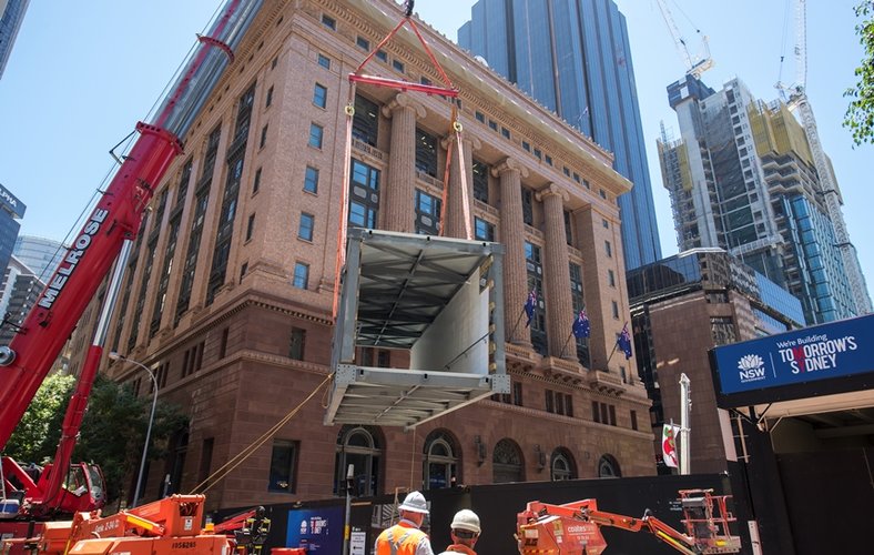 An on the ground view looking up at the pedestrian walkway being craned into place at the construction site of Sydney Metro's Martin Place Station. 