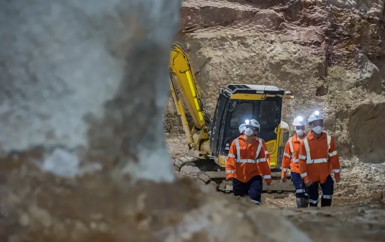 A close up shot of four construction workers walking inside a construction site with head torches shining bright inside with an excavator in the background at Sydney Metro's Martin Place Station.
