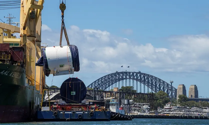 An on the ground view looking across at Tunnel Boring Machine being lifted off the boat by a large crane onto a smaller boat as it is arriving at Sydney Metro's The Bays construction site with shots of the Sydney Harbour Bridge in the background.