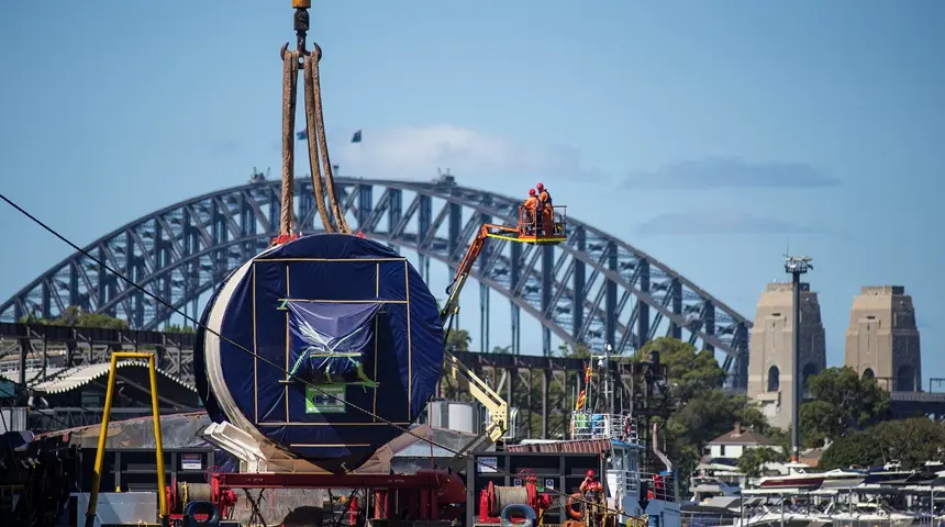 An on the ground view looking across at Tunnel Boring Machine arriving at Sydney Metro's The Bays construction site with shots of the Sydney Harbour Bridge in the background.