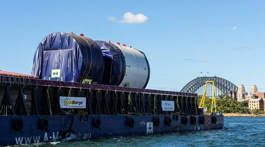 An on the ground view looking across at Tunnel Boring Machine on a boat as it is arriving at Sydney Metro's The Bays construction site with shots of the Sydney Harbour Bridge in the background.