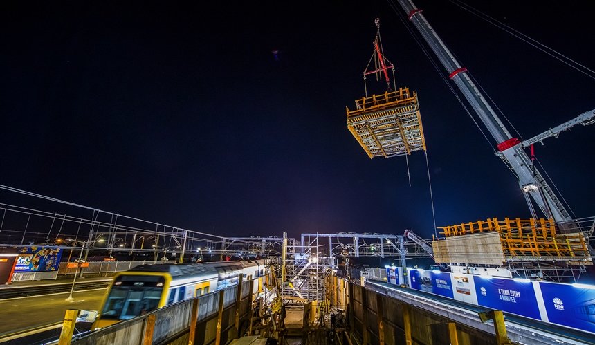 Frame work is lifted into place by a crane on the new concourse as it is being constructed at Sydney Metro's Sydenham Station. The is a train parked in the platform adjacent to the construction site at night time. 