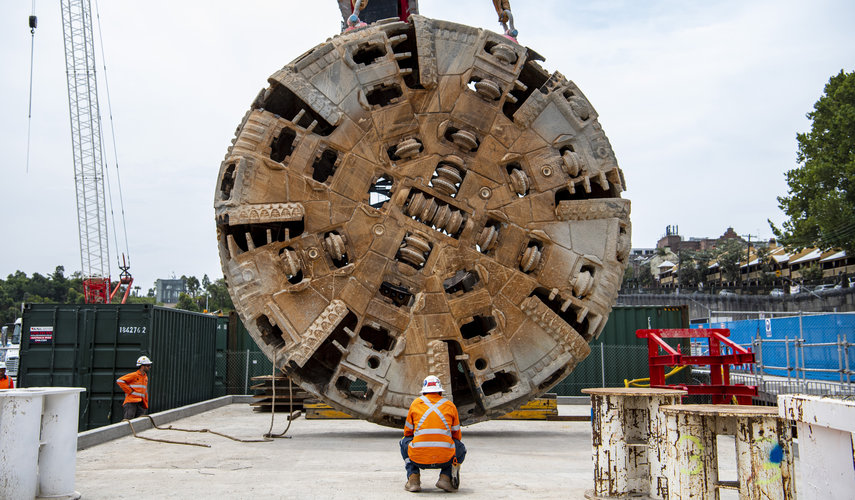 The cutterhead of tunnel boring machine Mum Shirl being lifted from the ground into the air by crane, whilst a construction worker supervises.