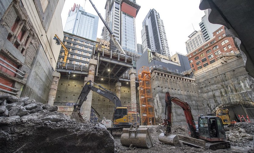 An on ground view looking up at heavy vehicle machinery working inside the Pitt Street South construction site. Tall buildings can be seen in the background.