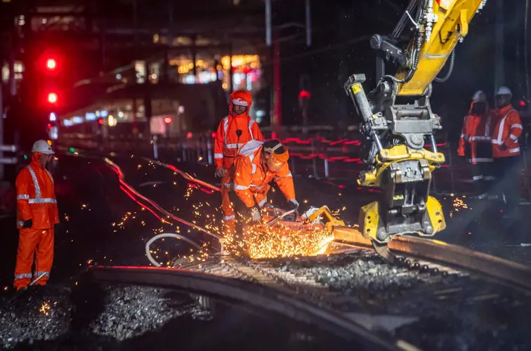 Machine cutting the train track at night. There is sparks coming off the track and 3 workers in personal protective equipment