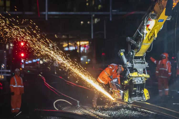 Machine cutting the train track at night. There is sparks coming off the track and 3 workers in personal protective equipment