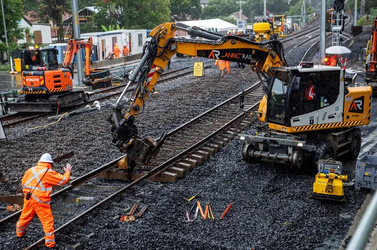 A heavy machine is picking up and moving the rail track. Construction worker in high viz is directing the operator of the machine.