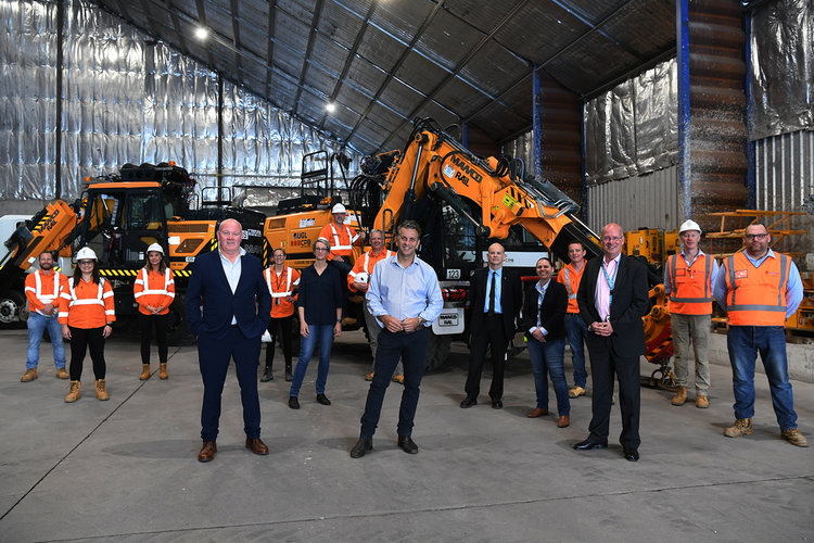 Construction workers and Sydney Metro executives pose for a photo at a construction site with heavy machinery in the background.
