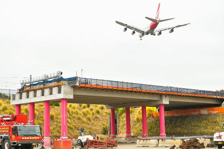 An aqueduct in Sydenham is lifted with a girder lift and sits on pink poles during construction while a plane flies overhead.