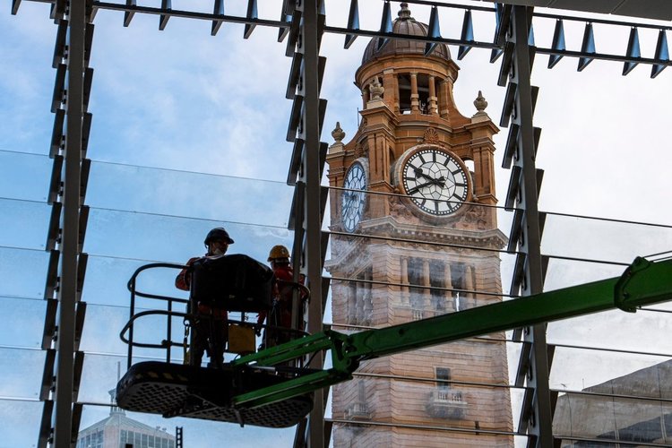 Two workers in high viz are standing on a cherry picker machine, with glass panes of a window behind them. The Central Station clock tower can be seen in the background.