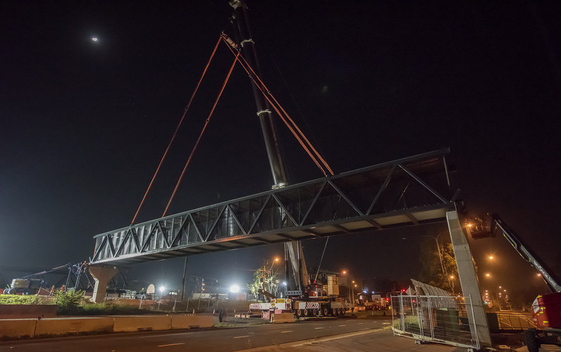 Construction on the Bella Vista pedestrian bridge at night time. The bridge crosses over a large road with many trucks and machinery on site.