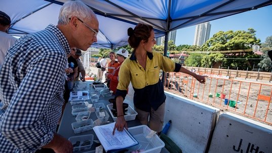 Sydney Metro employees showing community members the archaeological artefacts found at Waterloo Station pointing at a list of artefacts and where on the construction site behind them they were found. 