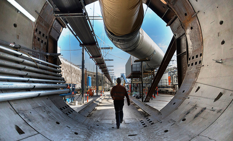 An on the ground view of a construction worker walking through Tunnel Boring Machine (TBM) 2 Florence with a large pipe overhead and construction site in the background. 