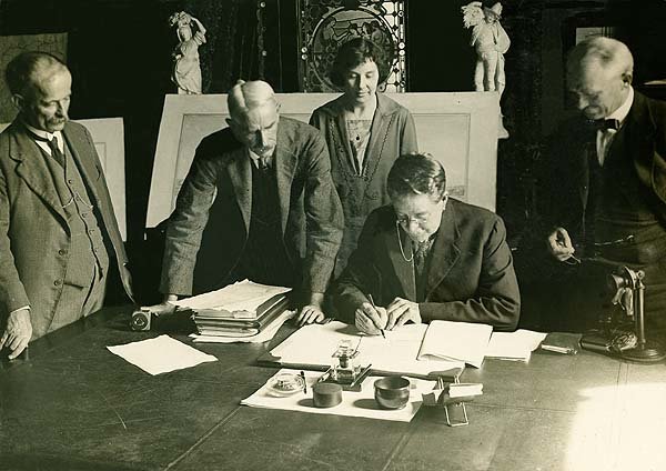 An  old photograph of Katheen Butler signing a document with four others gathers around her behind the table. 