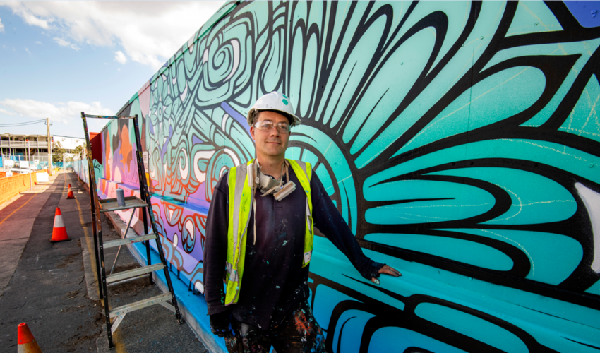 An artist in a high-vis vest and hard hat poses with the mural he has painted at Marrickville Metro Station. A ladder with a spray can on top sits behind him.