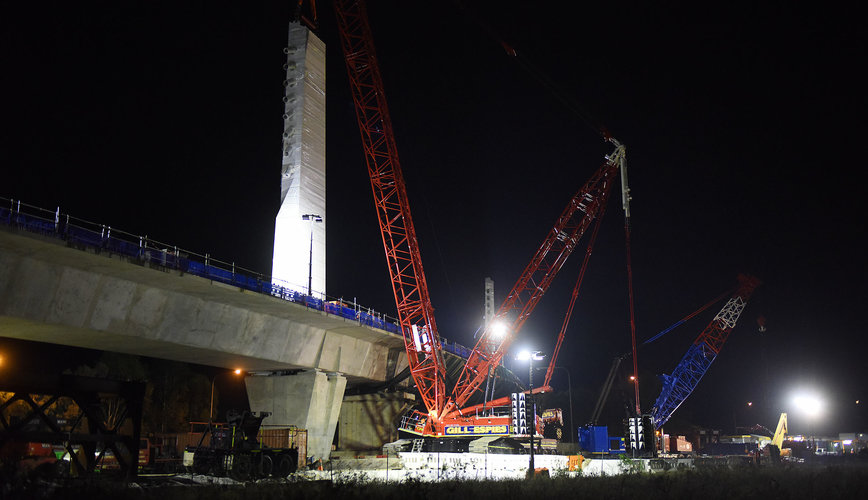 An on the ground view looking at the construction of the skytrain bridge tower as a crane lift is lifting material to the top of the tower at night at Sydney Metro's Rouse Hill. 