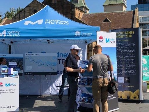 Close up shot showing the Sydney Metro community information stand  with a branded marquee, alignment maps, brochures on display and pull up banners as a Sydney Metro employee is talking to a member of the community at Parramatta Farmers Markets.