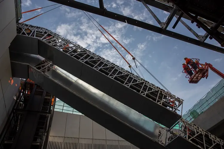 An on the ground view looking up as the double escalators are being crane lifted into place and two construction workers on a crane lift are inspecting the installation at Sydney Metro's Norwest site n May 2018.