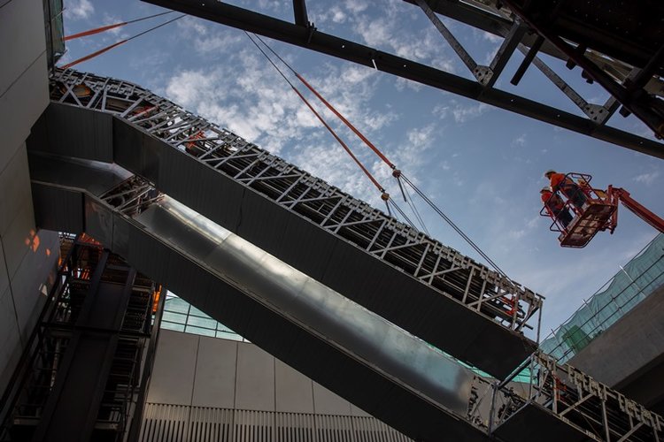 An on the ground view looking up as the double escalators are being crane lifted into place and two construction workers on a crane lift are inspecting the installation at Sydney Metro's Norwest site n May 2018. 