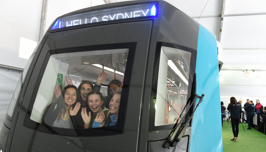 A group of five girls smiling and waving from the front of the Sydney Metro train carriage that was displayed at the Royal Easter Show. 
