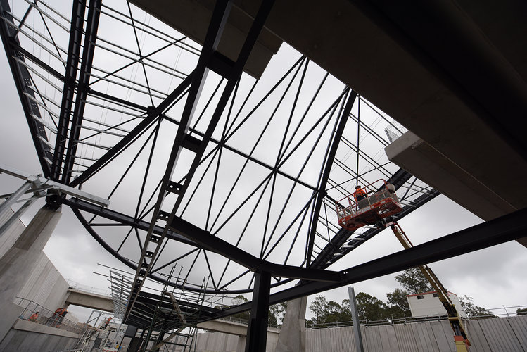 An on the ground view looking up at the steel beam reinforcement for the platform canopy at Sydney Metro's Cudgegong Road Station with a construction worker on a cherry picker carrying out work above. 