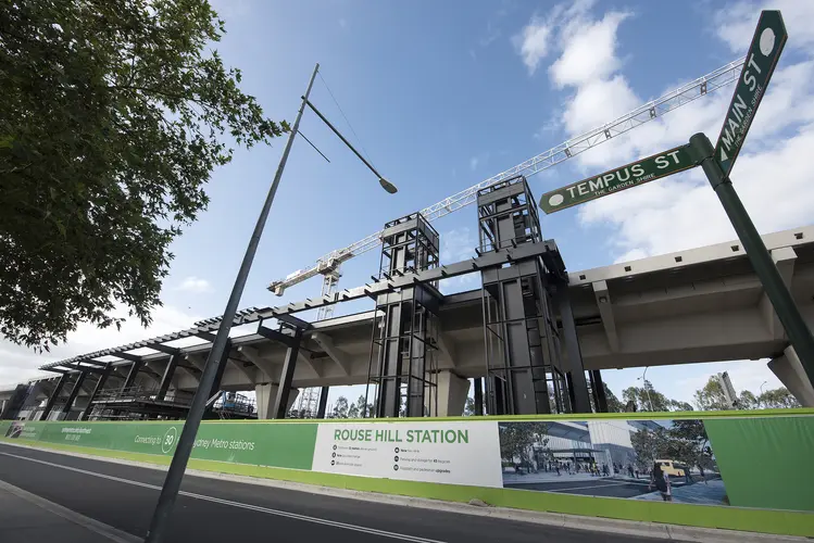 An on the ground view looking up at the station construction bridge with a large white crane in the background at Sydney Metro's new Rouse Hill Station from the corner of Tempus Street and Main Street.