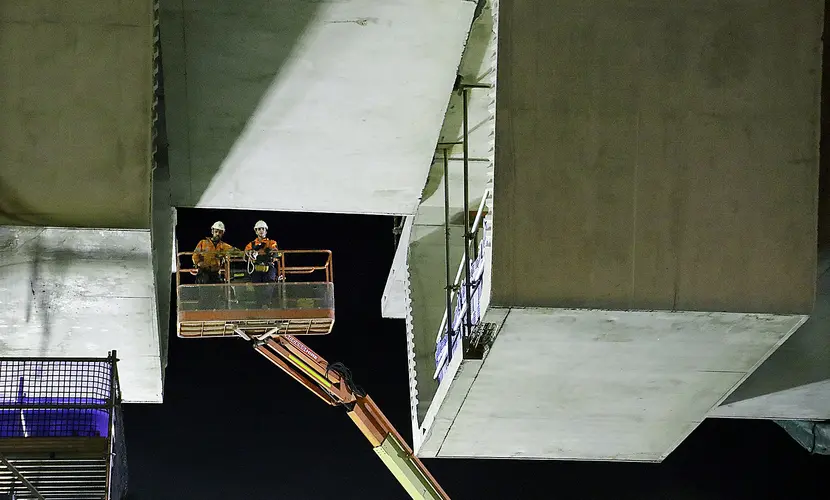 An on the ground view looking up at two construction workers on a crane lift as the final segment is nearing completion.