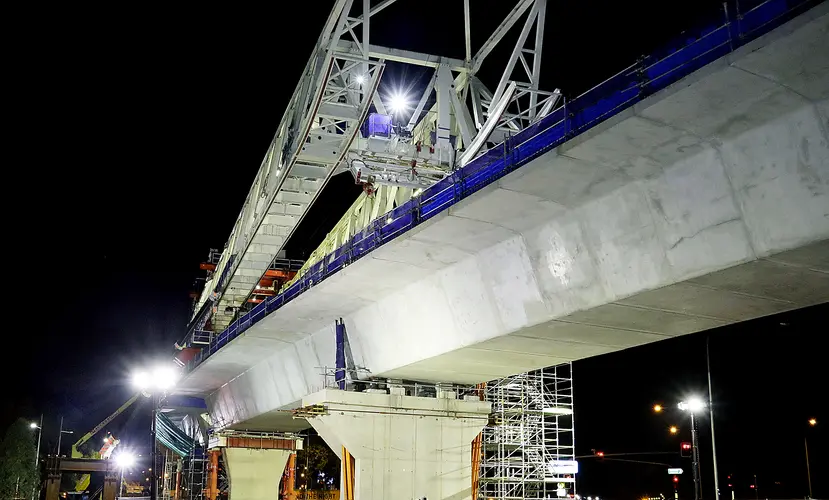 An on the ground view looking up at the construction work of the final span being lifted into place by a gantry crane at Sydney Metro's Rouse Hill Drive site.