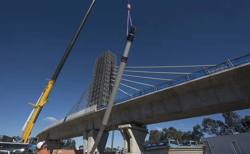 An on the ground view looking up at the construction of the railway bridge as cable poles are being lifted into place by a crane with the bridge tower in the background.