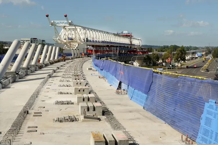 An on the ground view looking across at the construction of the railway bridge deck surface with rows of concrete slabs lined up to the right, pillars of concrete to the left and the large white gantry crane in the background.