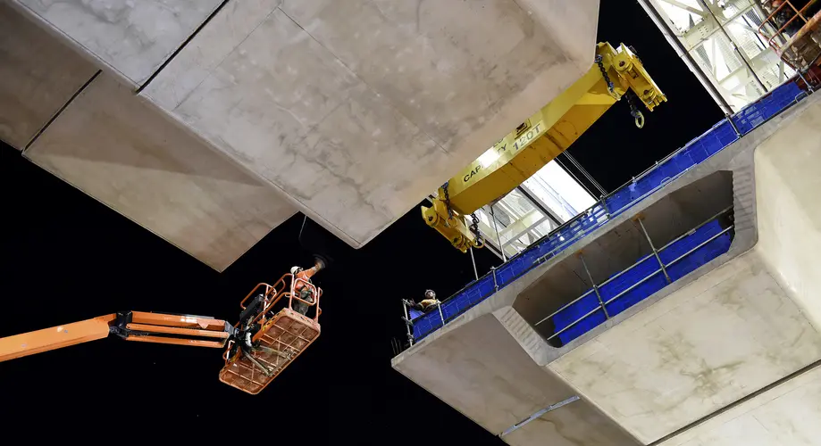 An on the ground shot looking up as the final segment of the Samantha Riley Drive bridge is being lifted into position with two construction workers guiding it in on a crane lift.
