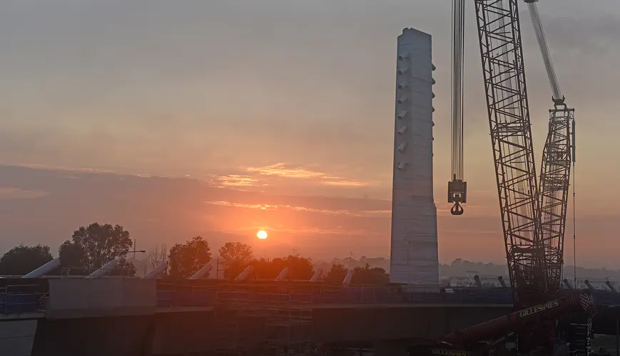 Looking across to the construction of Sydney Metro's skytrain bridge at sunrise. The image depicts a crane in the foreground with the bridge tower in the background.
