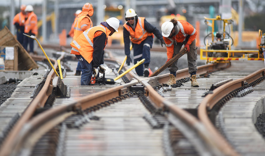 A group of four construction workers using machinery to lay the train tracks at a cross section, with more constructions completing the same task in the background. 