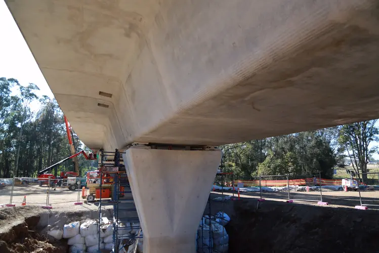A close up view under the bridge as construction work continues on Sydney Metro's skytrain platform, there are three heavy machinery in the background behind a fence of the platform construction. 