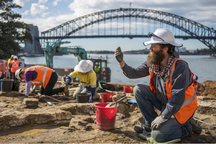 Archaeologists in high vis and hard hats are on their hands and knees while working in the excavation area at the Blues Point construction site. Sydney Harbour Bridge is seen in the background.