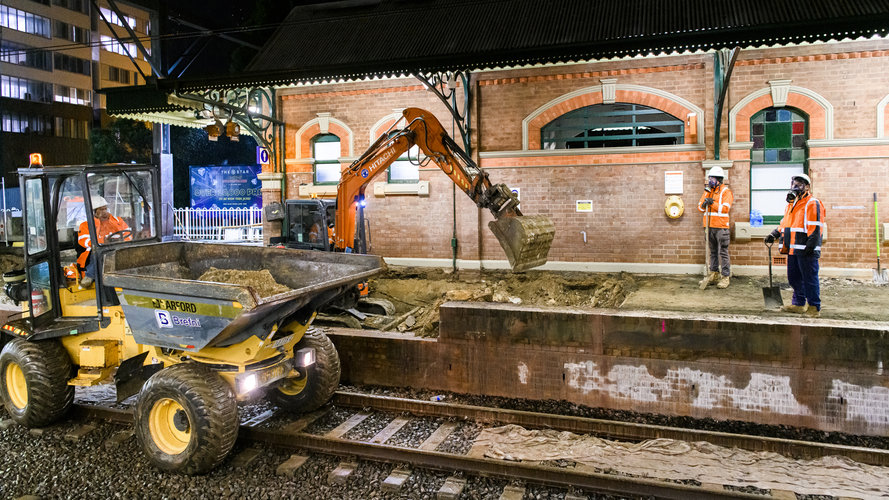 Two construction workers in orange high-vis watch an excavator digging while performing track works at Canterbury Station.