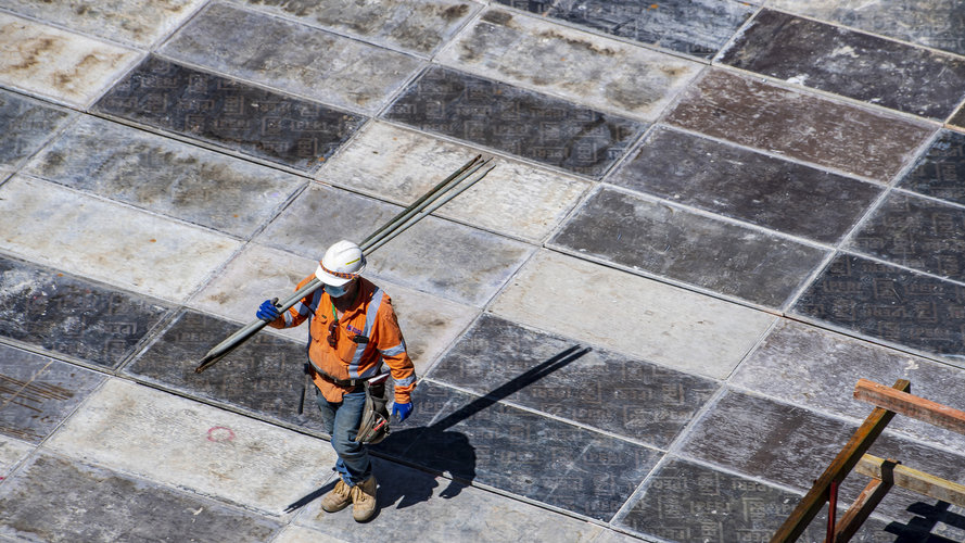 A birds eye view of a construction worker walking with metal rods over his shoulder.