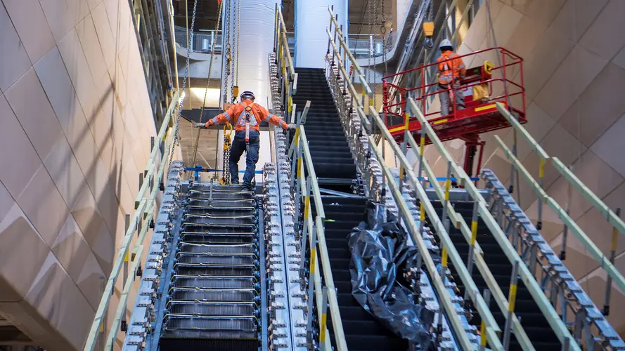 One construction worker stands at the top installing escalators at Central Station while another is in a lift next to him.