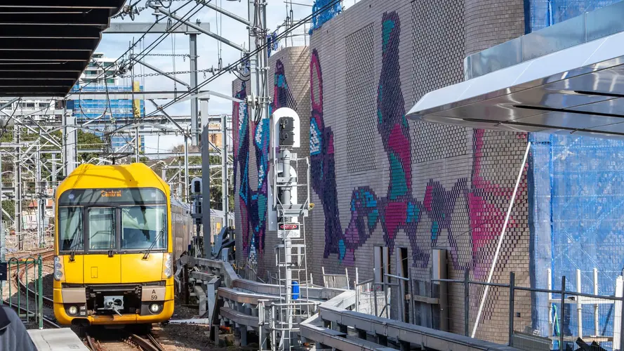 Colourful aboriginal artwork covers the brick ventilation buildings along the tracks at Central Station while a yellow train is approaching the platform.