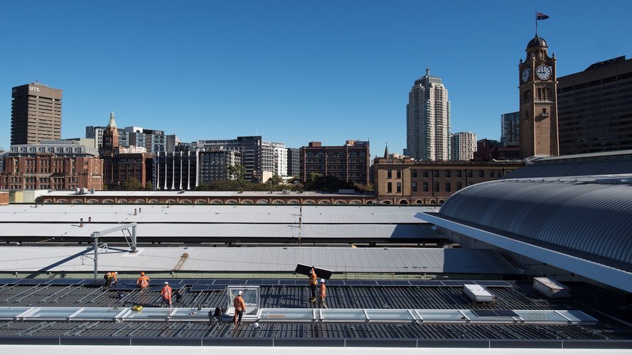 Wide shot of construction workers installing solar panels on Central Station's rooftop with Sydney's skyline in the background